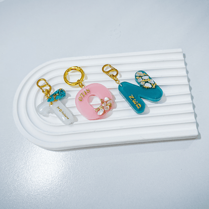 White letter T personalised initial custom pet ID tag with gold-plated stainless steel hardware, turquoise blue stones and gold flake, pink letter O pet ID tag with seashells and gold flake, blue letter N pet ID tag with seashells and gold flake..